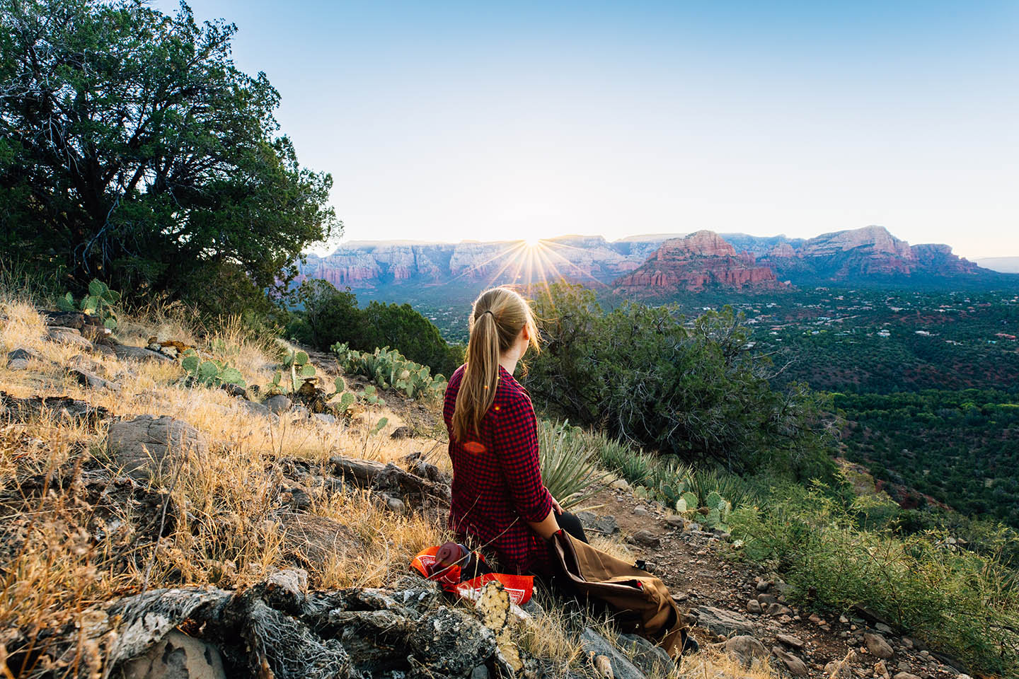 a woman sits on a hilltop looking away at the sun rising over the mesa